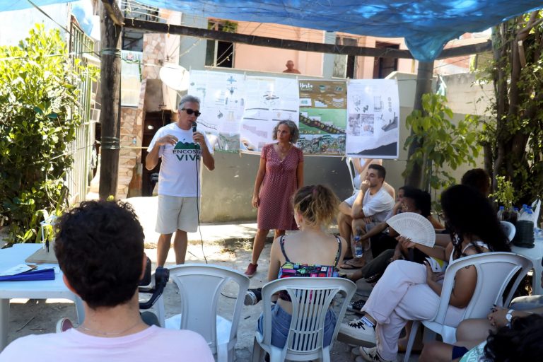 Apresentação ao ar livre numa comunidade: homem fala ao microfone ao lado de uma mulher, com painéis de projeto pendurados atrás e moradores sentados em cadeiras de plástico sob uma lona azul