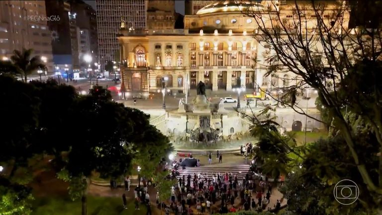 Vista noturna do Vale do Anhangabaú, em São Paulo, com o Theatro Municipal iluminado ao fundo, o monumento e a fonte ao centro e público reunido na escadaria