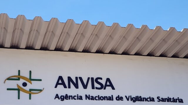 Fachada da sede da Anvisa em Brasília com a placa da agência e o logotipo sobre fundo branco