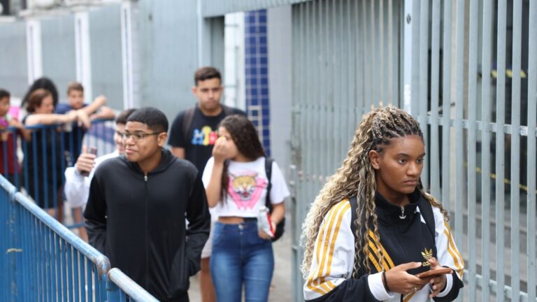 Jovens caminham em frente ao portão gradeado de uma escola onde o Enem é aplicado