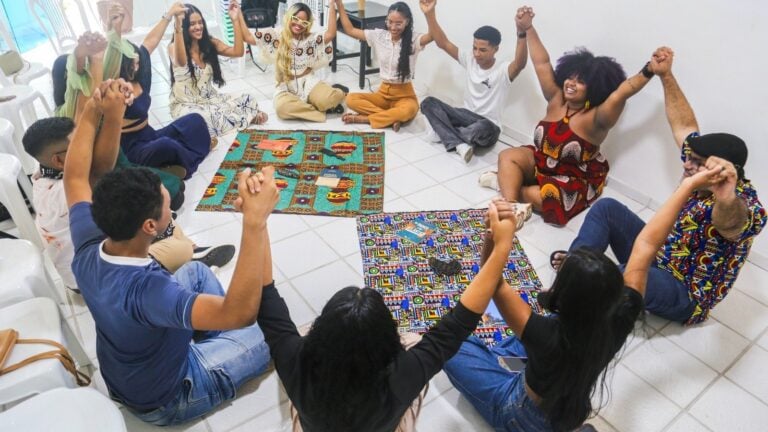 Jovens sentados em círculo no chão, de mãos dadas e sorrindo, sobre tapetes com estampas africanas