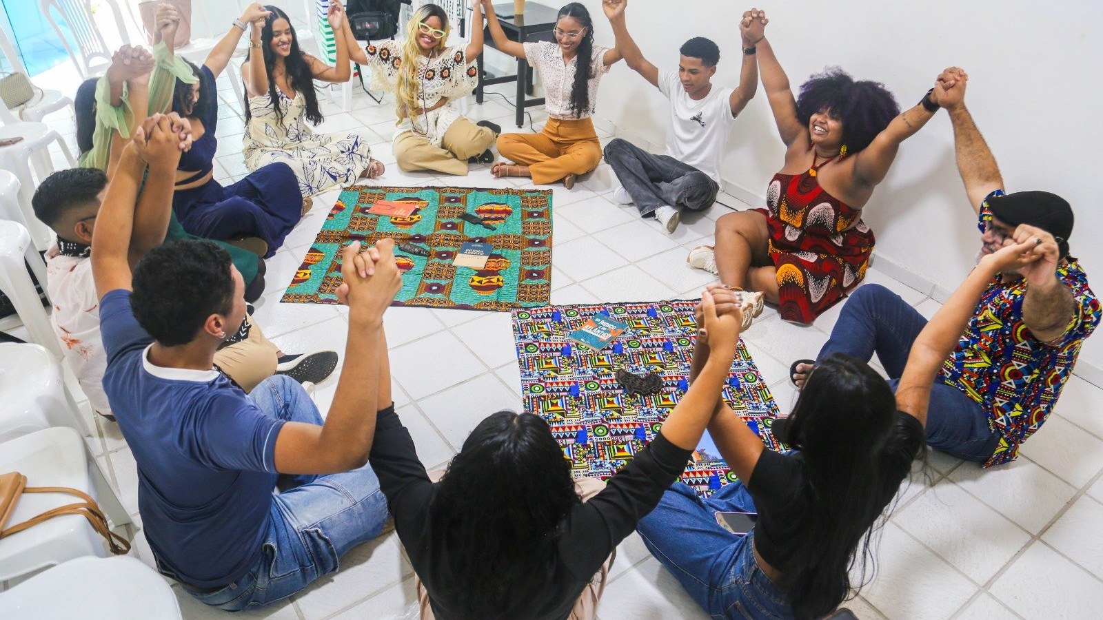 Jovens sentados em círculo no chão, de mãos dadas e sorrindo, sobre tapetes com estampas africanas