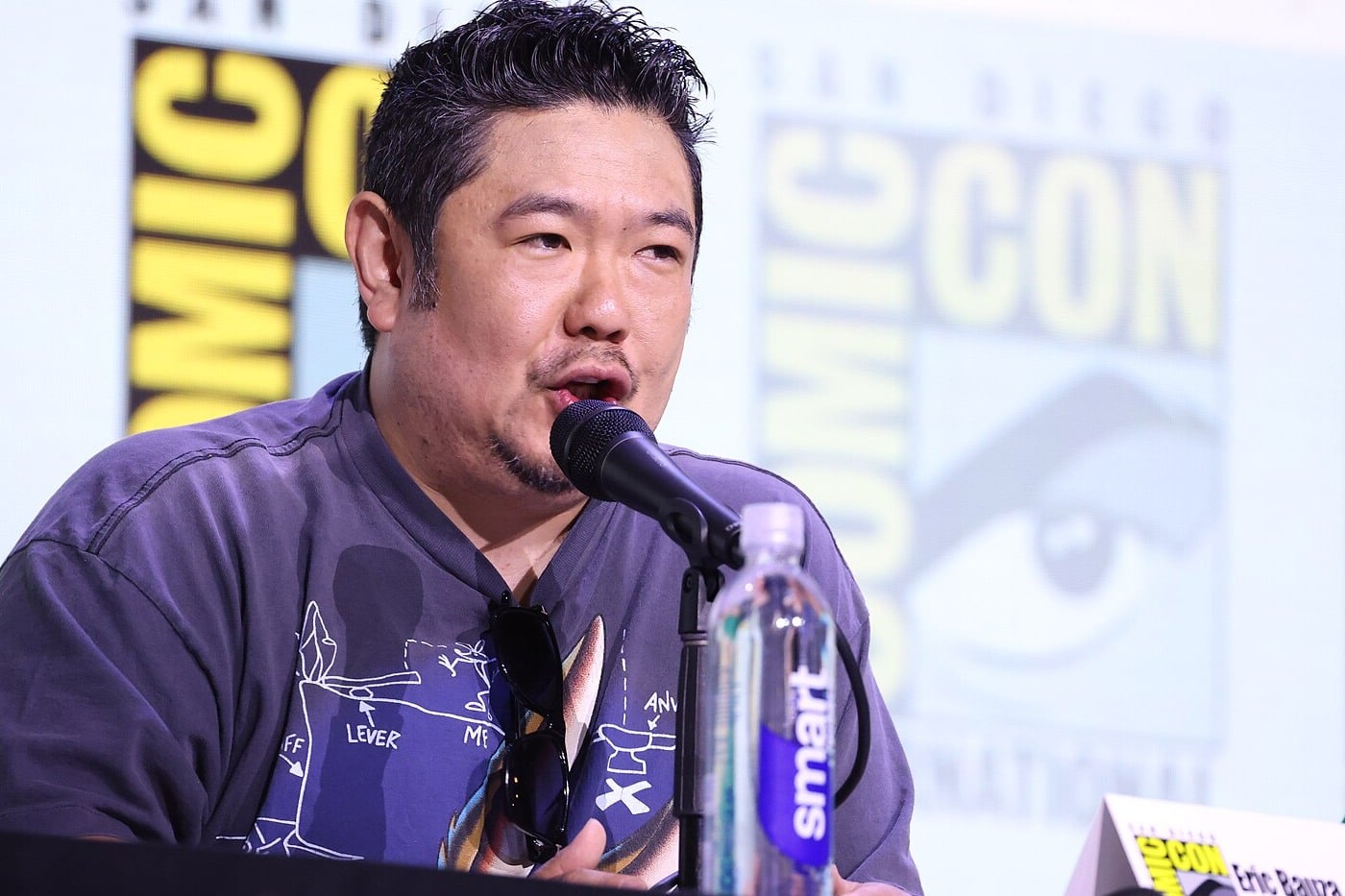 Eric Bauza fala ao microfone durante painel, com o logotipo da San Diego Comic-Con ao fundo