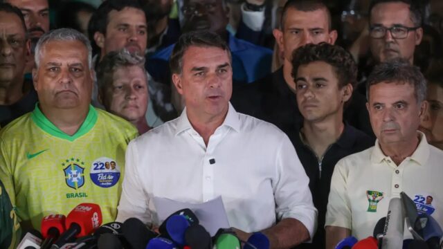 Flávio Bolsonaro, de camisa branca, fala diante de um conjunto de microfones, cercado por apoiadores de camisa amarela da campanha