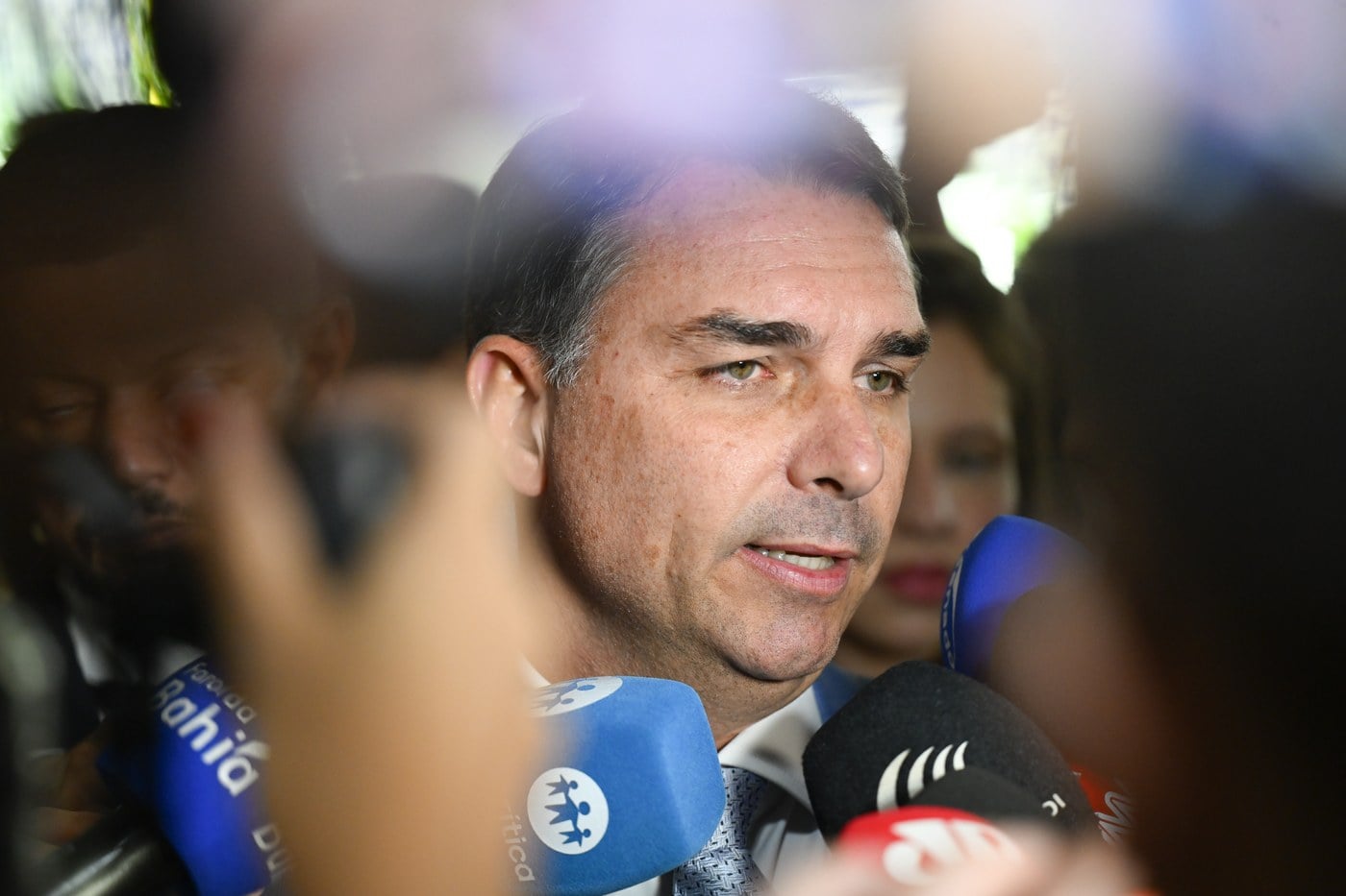 Flávio Bolsonaro, de terno azul-claro, fala cercado por microfones de emissoras de rádio e televisão