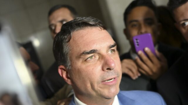 Flávio Bolsonaro, de terno azul, cercado por microfones e celulares de jornalistas