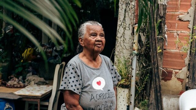 Mulher idosa em pé no quintal de uma casa, cercada por plantas, usando blusa cinza com estampa