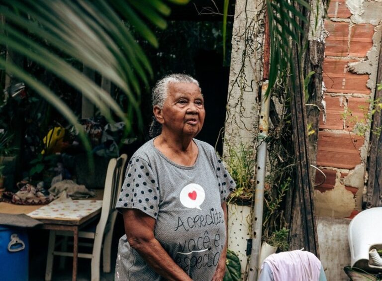 Mulher idosa em pé no quintal de uma casa, cercada por plantas, usando blusa cinza com estampa
