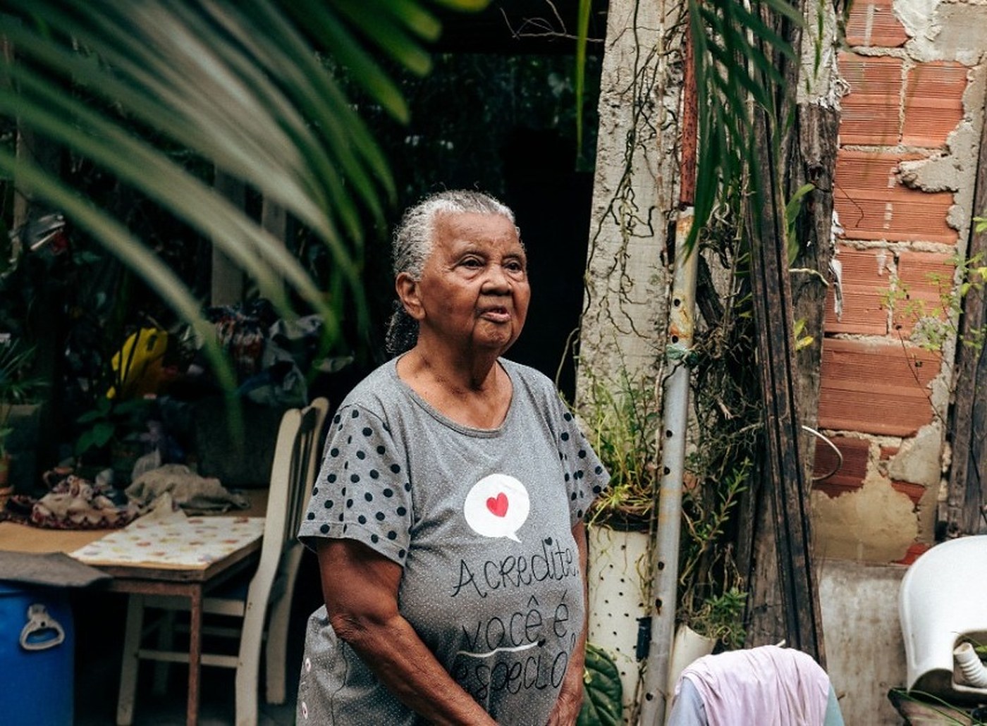 Mulher idosa em pé no quintal de uma casa, cercada por plantas, usando blusa cinza com estampa