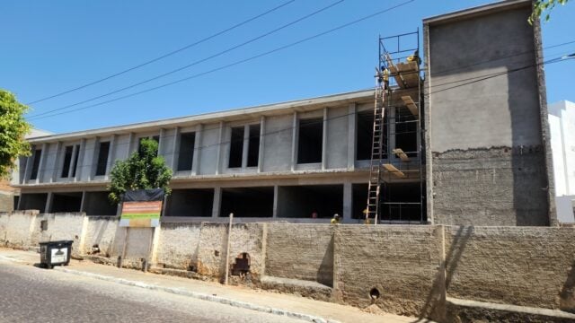 Fachada de escola de dois pavimentos em construção, com andaimes e trabalhadores na obra
