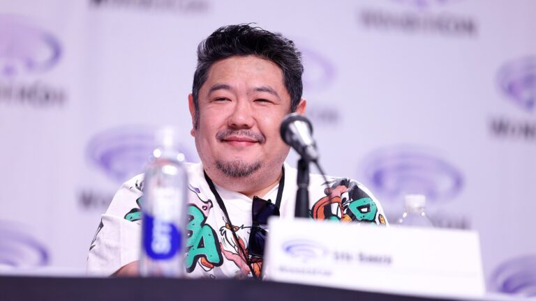 Eric Bauza sorri diante de um microfone, com o logotipo da WonderCon ao fundo
