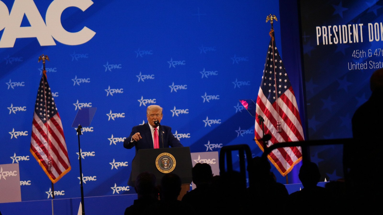 Donald Trump discursa ao pódio diante de bandeiras dos Estados Unidos, com a plateia em silhueta