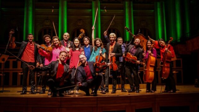 Grupo de músicos de cordas posa no palco com violinos, violas, cellos e um contrabaixo
