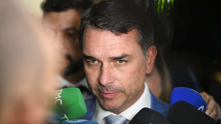 Flávio Bolsonaro, de terno azul, cercado por microfones de emissoras durante entrevista no Senado