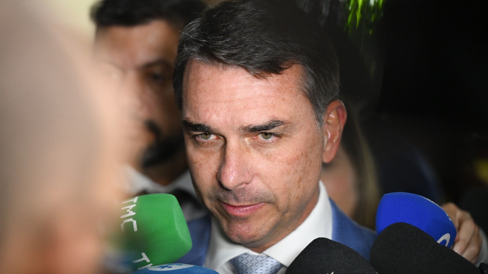 Flávio Bolsonaro, de terno azul, cercado por microfones de emissoras durante entrevista no Senado