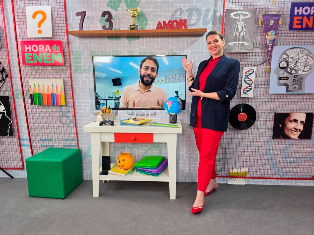 Apresentadora ao lado de monitor com professor e da placa Hora do Enem, em estúdio de TV