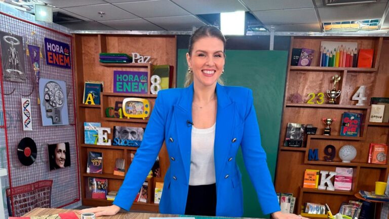 Apresentadora de blazer azul sorri diante de estante com livros e da placa Hora do Enem