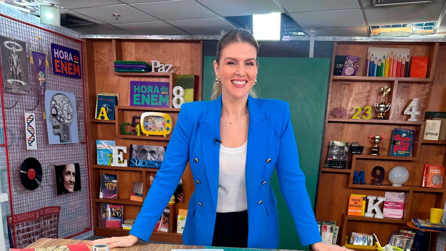 Apresentadora de blazer azul sorri diante de estante com livros e da placa Hora do Enem