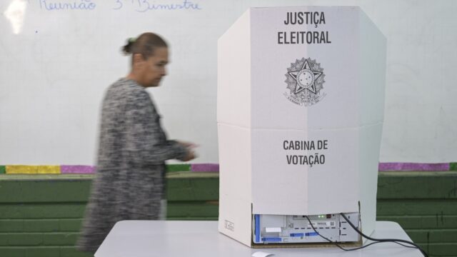 Cabina de votação com a urna eletrônica sobre uma mesa em sala de aula, com uma pessoa passando ao fundo