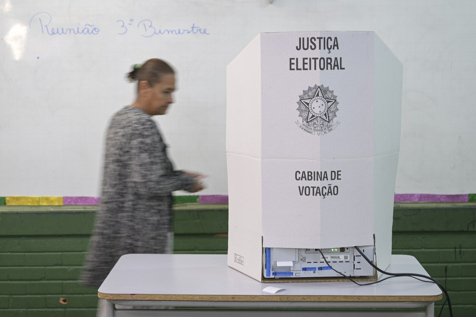 Cabina de votação com a urna eletrônica sobre uma mesa em sala de aula, com uma pessoa passando ao fundo
