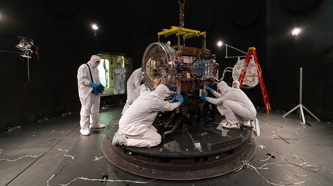 Técnicos de macacão branco instalam a nave LINK, com três braços robóticos, sobre uma base dentro de uma câmara escura de testes
