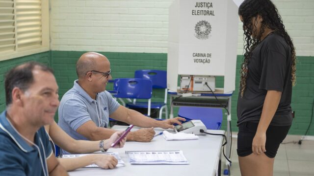 Dois mesários sentados à mesa atendem uma eleitora em pé, ao lado da urna eletrônica e da cabina de votação