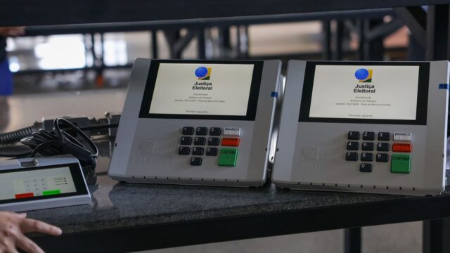 Duas urnas eletrônicas sobre uma bancada, com a tela inicializando o software de votação, ao lado de um terminal do mesário