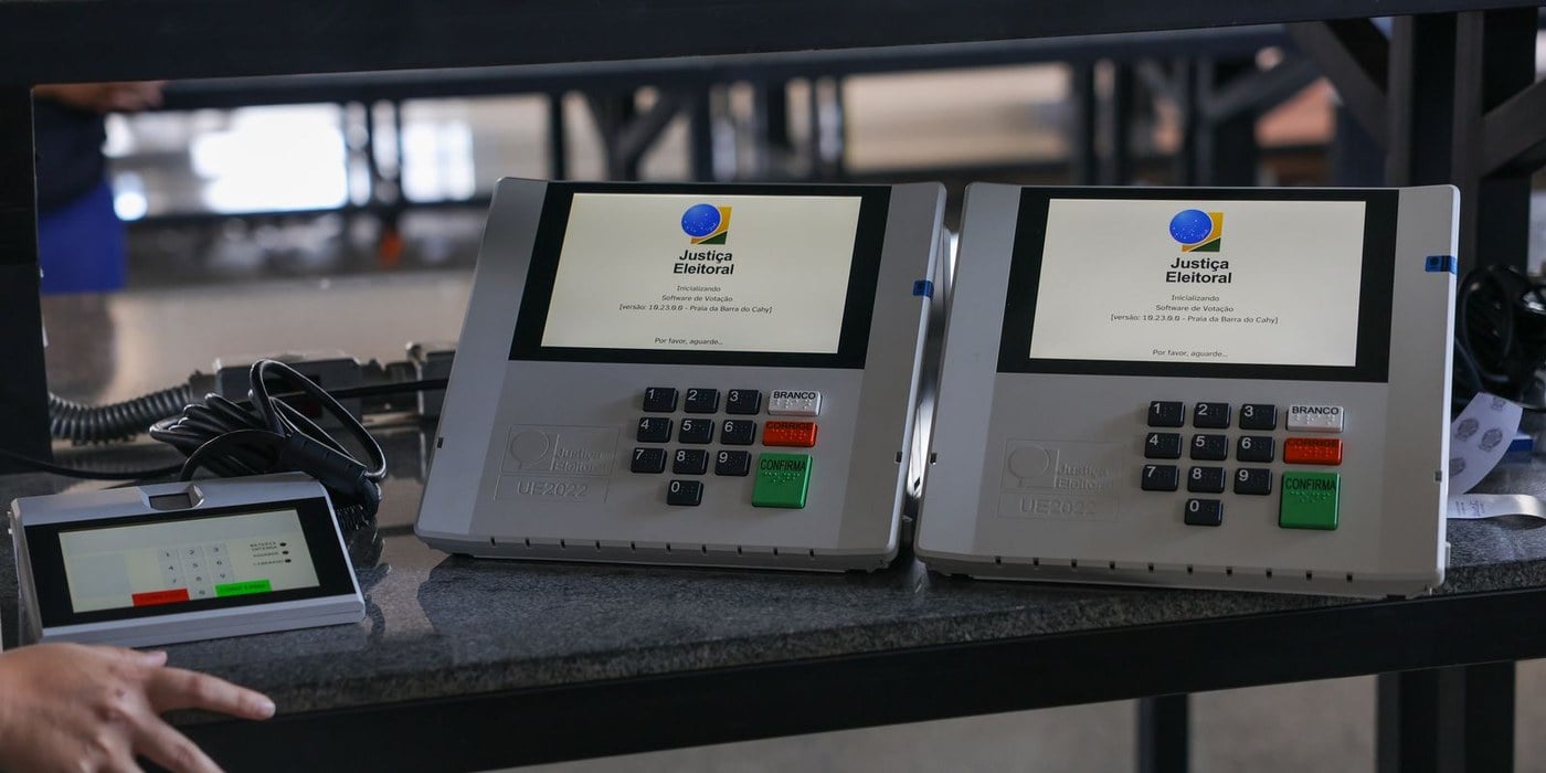 Duas urnas eletrônicas sobre uma bancada, com a tela inicializando o software de votação, ao lado de um terminal do mesário