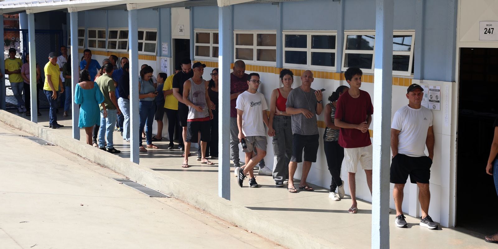 Fila de eleitores ao lado de escola pública com placa de seção eleitoral, no segundo turno das eleições de 2026